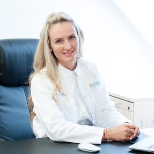 ELFIN Care Geschäftsleitung Ksenija Pies ist zu sehen. Sie sitzt an ihrem Schreibtisch und lächelt in die Kamera. Sie hat laange blonde Haare und trägt einen weißen Kittel.