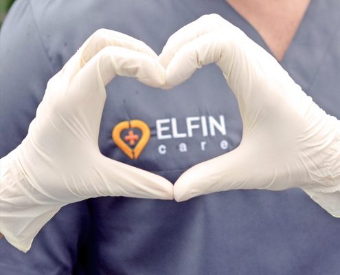 Person in medizinischer Berufskleidung formt mit beiden Händen, die mit weißen Einmalhandschuhen bedeckt sind, ein Herz. Auf der Kleidung ist das Logo von „ELFIN care“ mit einem Herzsymbol und einem Kreuz zu sehen.