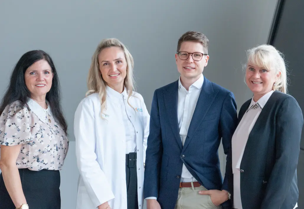 Drei Mitarbeiterinnen von Elfin Care stehen nebeneinander und lächeln in die Kamera. Eine trägt einen grauen Kittel mit Stethoskop, die zweite ein weißes Hemd mit Elfin Care Logo und Namensstickerei, die dritte einen türkisfarbenen Pflegekittel mit Logo. Der Hintergrund ist grau.