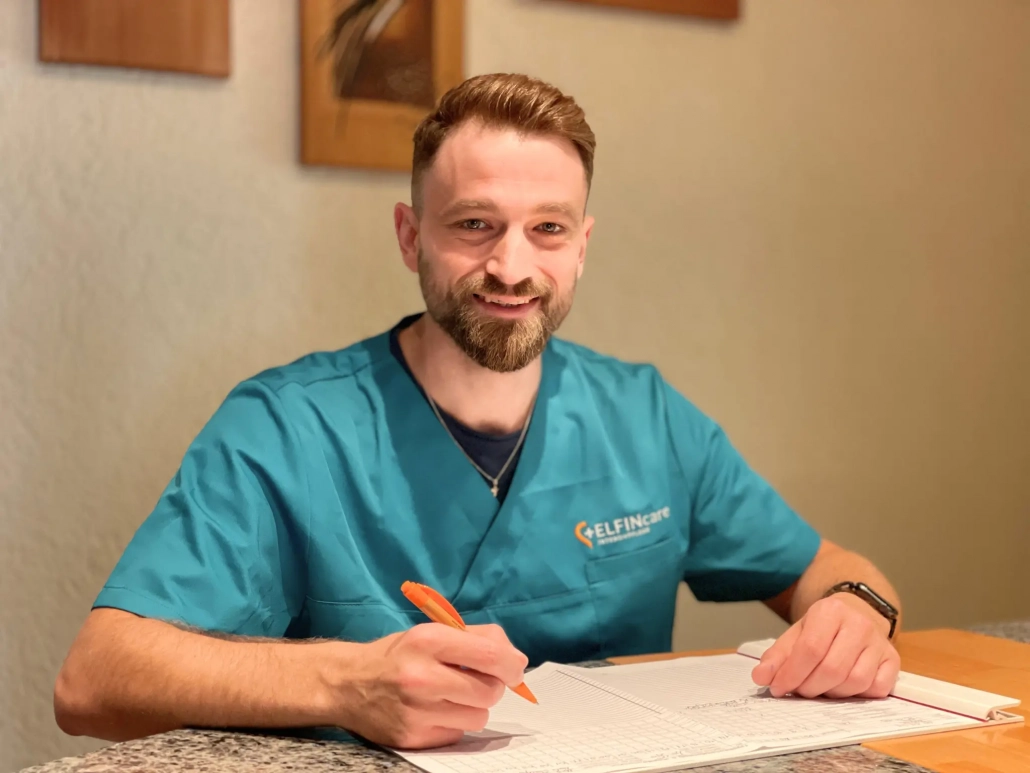 Ein Pfleger in türkisfarbener Arbeitskleidung mit Elfin Care Logo sitzt an einem Tisch, lächelt und schreibt mit einem Stift in ein Formular oder Notizbuch.