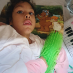 Ein Mädchen liegt auf einem Bett und hält ein großes grünes, stacheliges Spiel- oder Therapiegerät in den Händen. Sie trägt einen weißen Pullover mit rosa Ärmeln. Hinter ihrem Kopf liegt ein Kissen mit einer Zeichnung von zwei Comic-Tieren, einem Bären mit Fliegerbrille und einem Schaf mit orangefarbener Mütze. Rechts im Bild ist ein Teil eines Spielzeug-Keyboards zu sehen.
