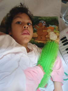 Ein Mädchen liegt auf einem Bett und hält ein großes grünes, stacheliges Spiel- oder Therapiegerät in den Händen. Sie trägt einen weißen Pullover mit rosa Ärmeln. Hinter ihrem Kopf liegt ein Kissen mit einer Zeichnung von zwei Comic-Tieren, einem Bären mit Fliegerbrille und einem Schaf mit orangefarbener Mütze. Rechts im Bild ist ein Teil eines Spielzeug-Keyboards zu sehen.
