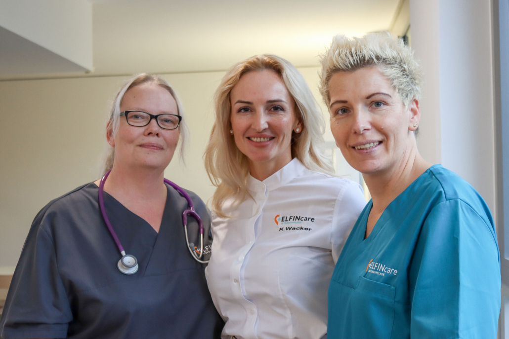 Drei Mitarbeiterinnen von Elfin Care stehen nebeneinander und lächeln in die Kamera. Eine trägt einen grauen Kittel mit Stethoskop, die zweite ein weißes Hemd mit Elfin Care Logo und Namensstickerei, die dritte einen türkisfarbenen Pflegekittel mit Logo.