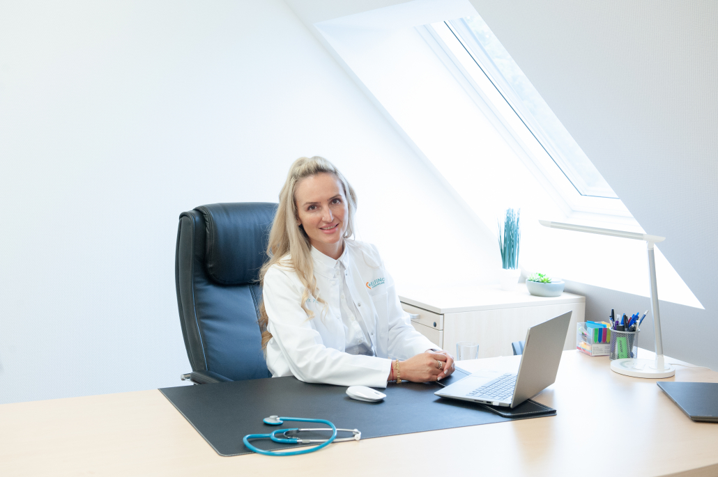 Eine Frau in weißem Kittel mit Elfin Care Logo sitzt an einem Schreibtisch vor einem Laptop. Auf dem Tisch liegen ein Stethoskop, eine Computermaus und Büroutensilien. Durch ein Dachfenster fällt Tageslicht ins moderne Büro.