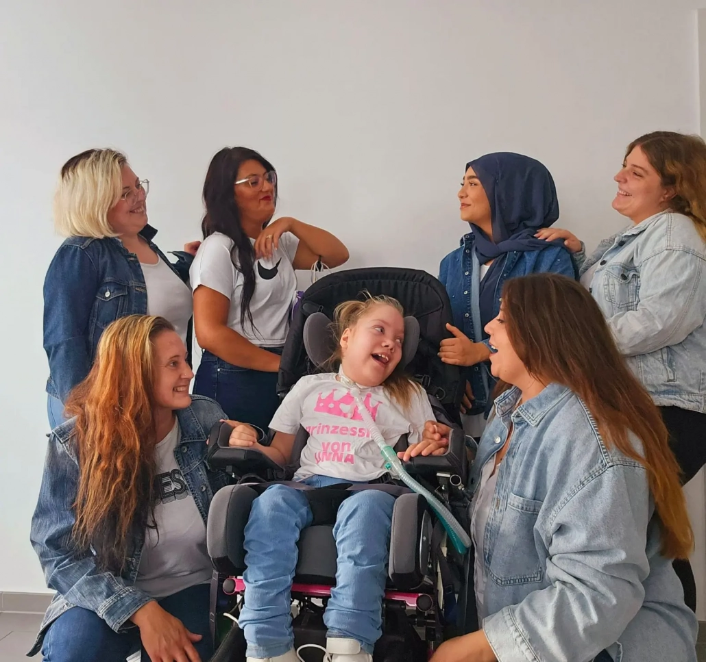 Eine Gruppe von sechs Frauen sitzt und steht um ein lächelndes Mädchen im Rollstuhl. Alle wirken fröhlich und lachen miteinander, einige tragen Jeansjacken. Das Mädchen trägt ein weißes T-Shirt mit rosa Krone und dem Aufdruck „Prinzessin von…“