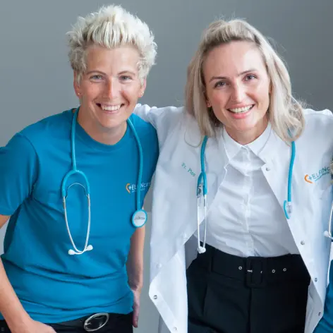 Team ELFIN Care Intensivpflege zu hause Ambulante Intensivpflege zu Hause: Zwei Pflegefachkräfte mit Stethoskopen lächeln in die Kamera und stehen eng nebeneinander, symbolisch für persönliche und professionelle Betreuung von Patientinnen und Patienten im häuslichen Umfeld.